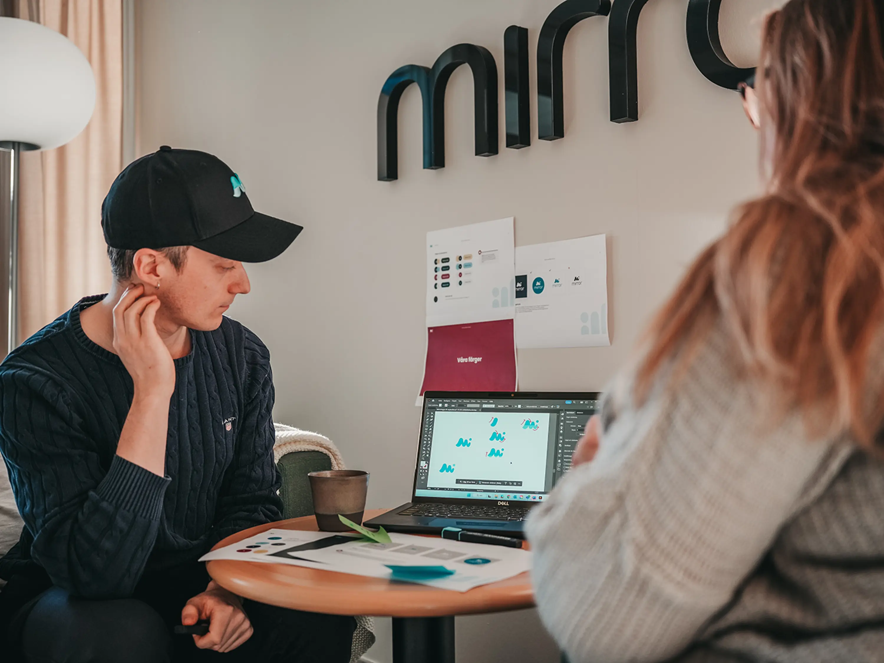 Två personer som sitter vid ett runt bord och tittar på en dator med designelement